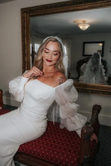 A woman in a white wedding dress with long sleeves and a veil, sitting on a red upholstered bench, smiling with closed eyes and hand near face in a room with a large mirror and a tattoo on her chest.