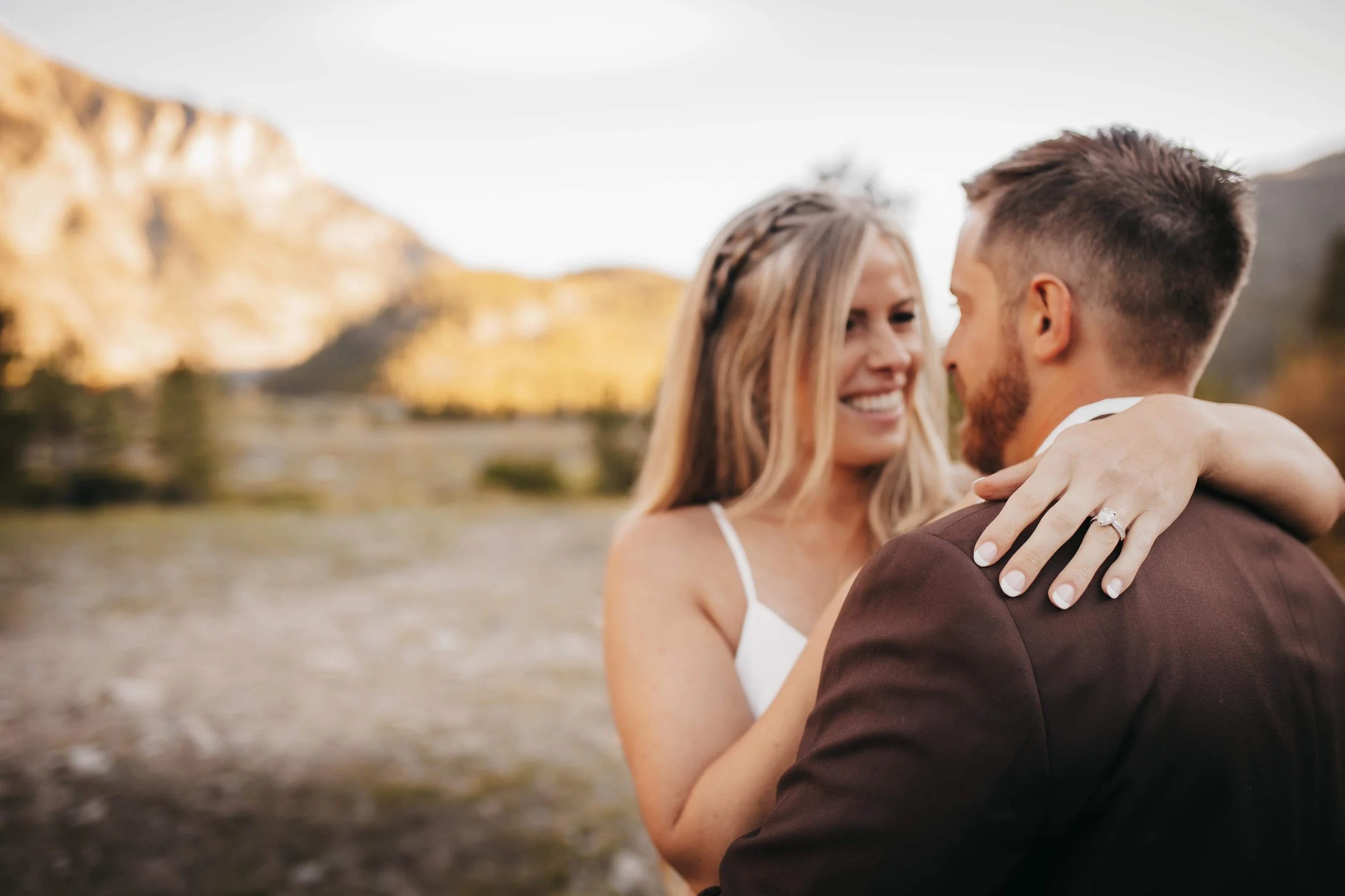A couple embracing outdoors near a mountain landscape, with the woman showing an engagement ring on her finger.
