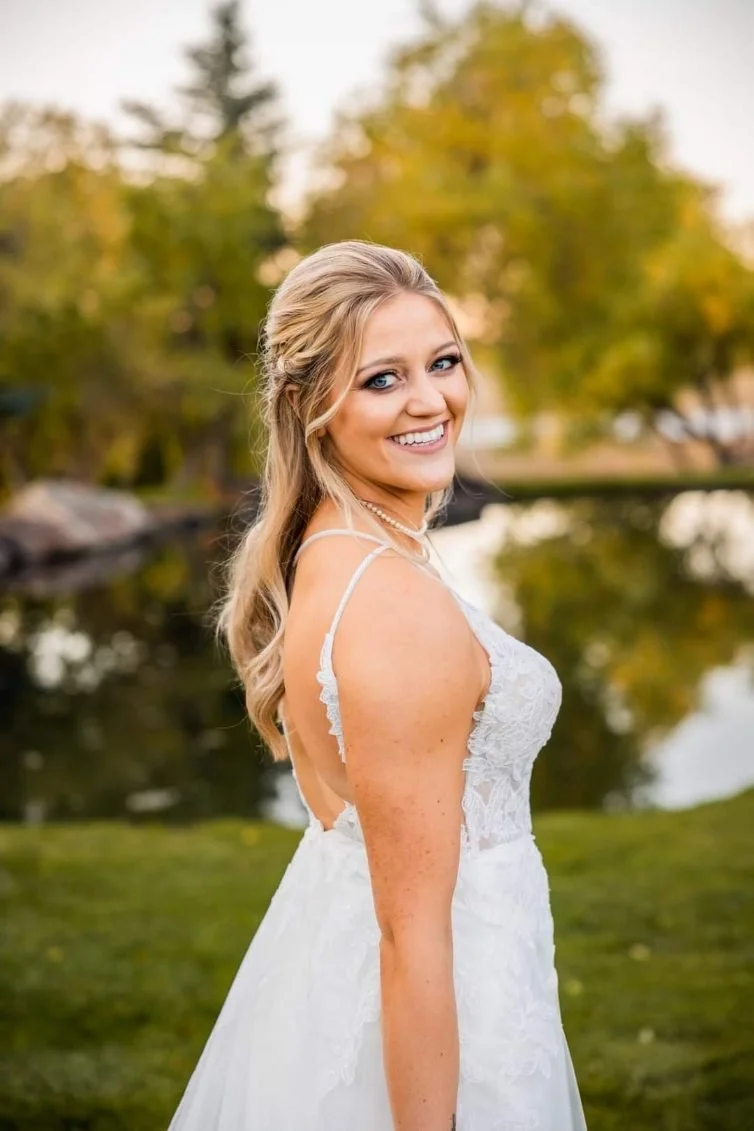 A smiling woman in a white lace dress standing outdoors near a body of water with trees in the background, during daytime.