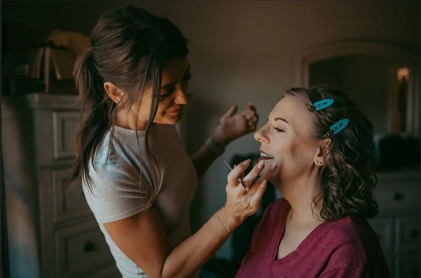 A woman with blue hair clips smiling as another woman applies lipstick to her lips. The second woman has dark, curly hair and is wearing a maroon top. They are indoors in a warmly lit room.