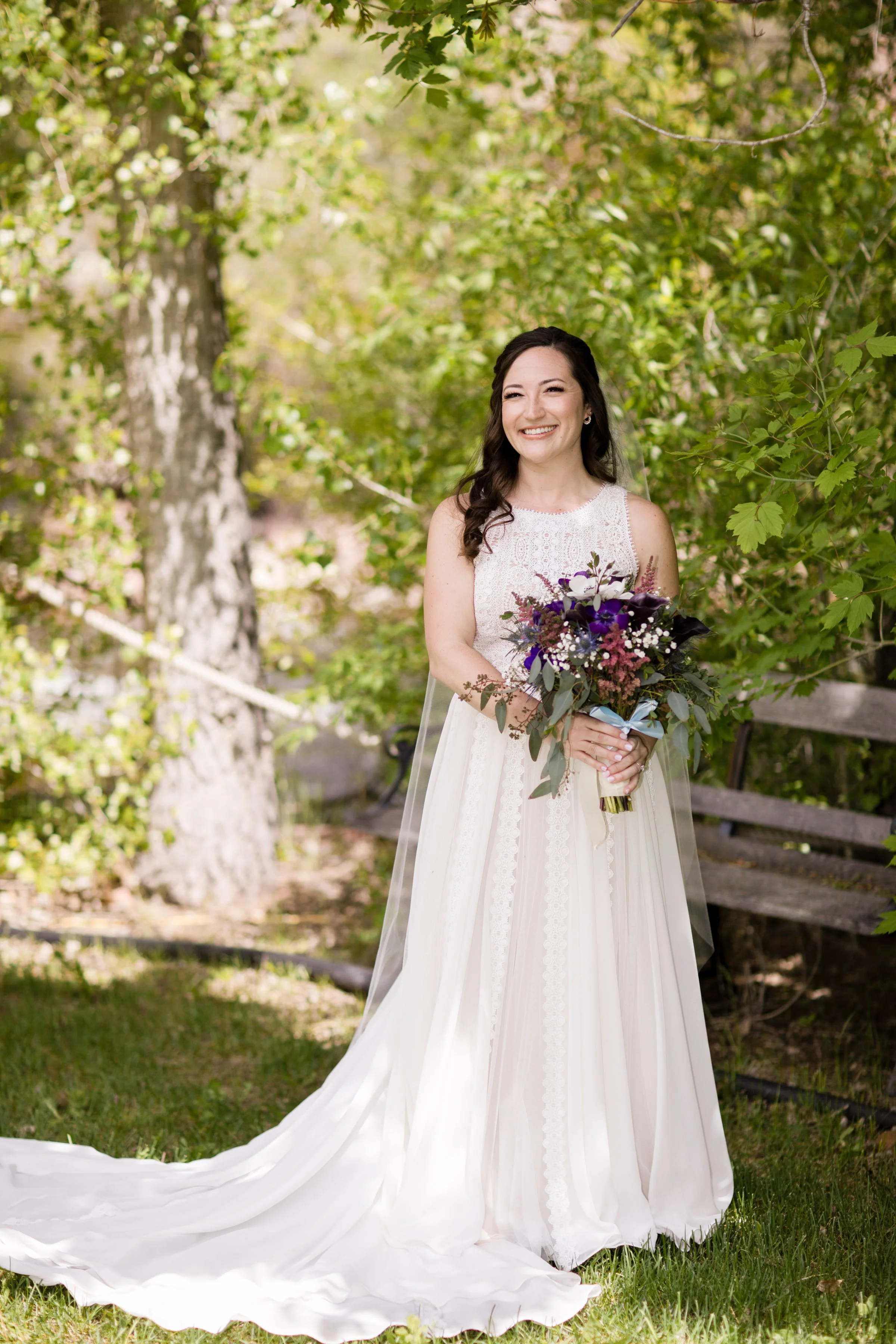 A woman in a wedding dress holding a bouquet of flowers, standing outdoors in a lush green area with trees and a wooden bench in the background.