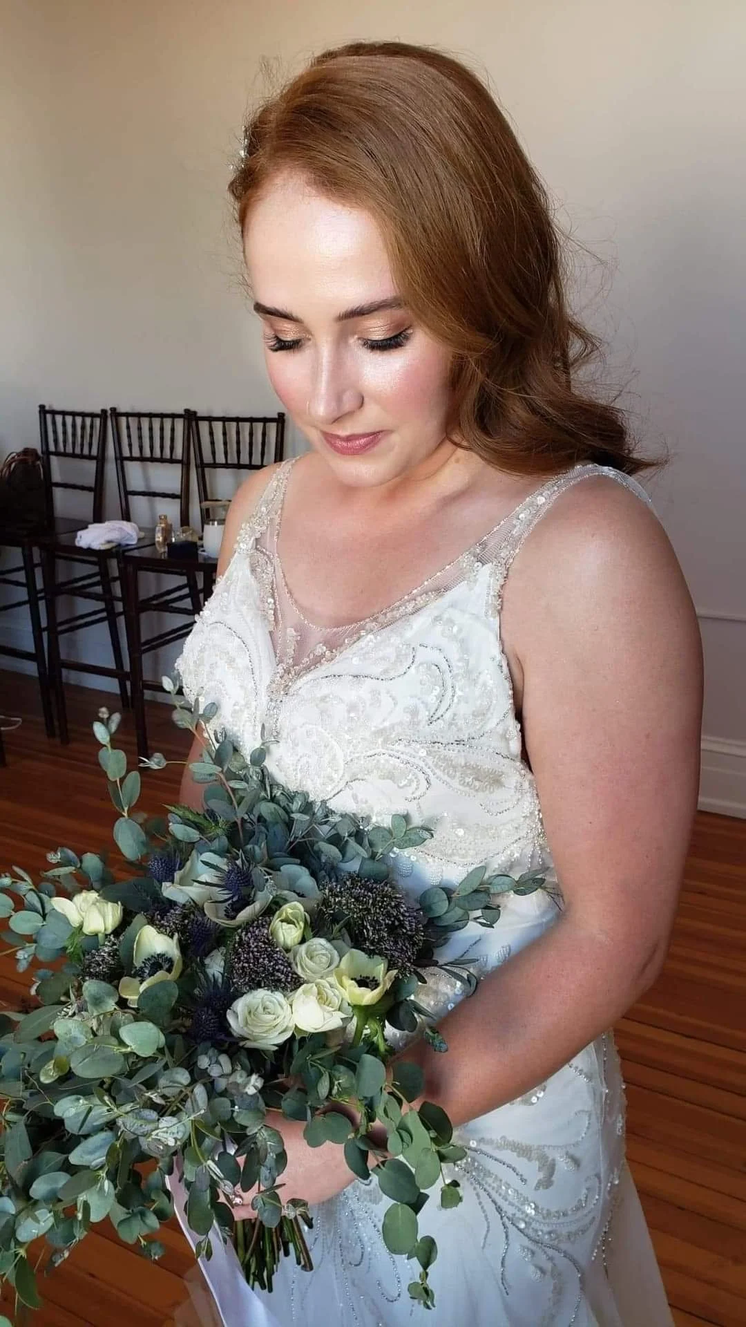 A red-haired woman in a white wedding dress holding a bouquet of flowers, standing indoors with wooden floors and chairs in the background.