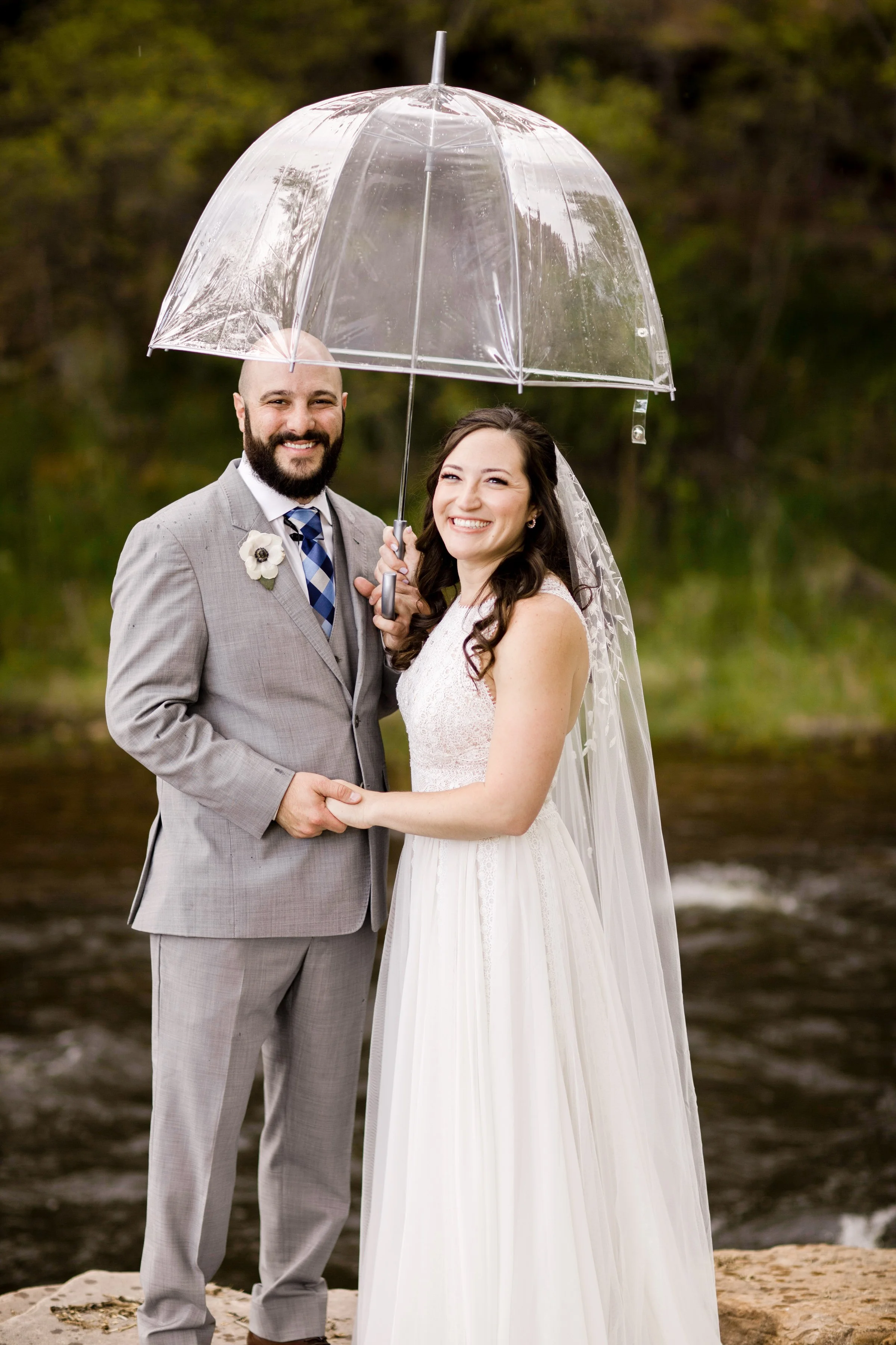 A smiling bride and groom holding hands and sharing an umbrella outdoors during their wedding, with a river and trees in the background.