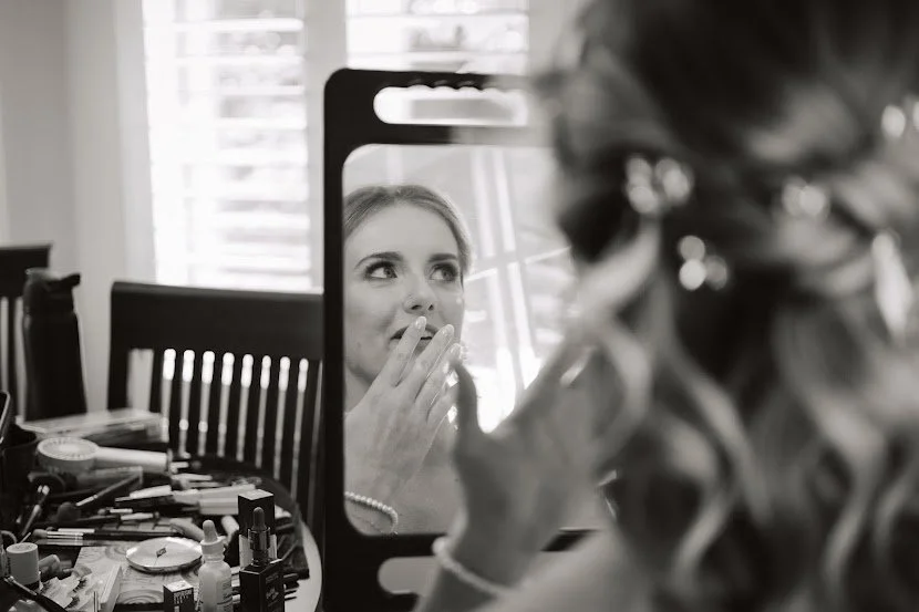 A woman applying makeup while looking in a mirror on a cluttered table with makeup products and brushes.