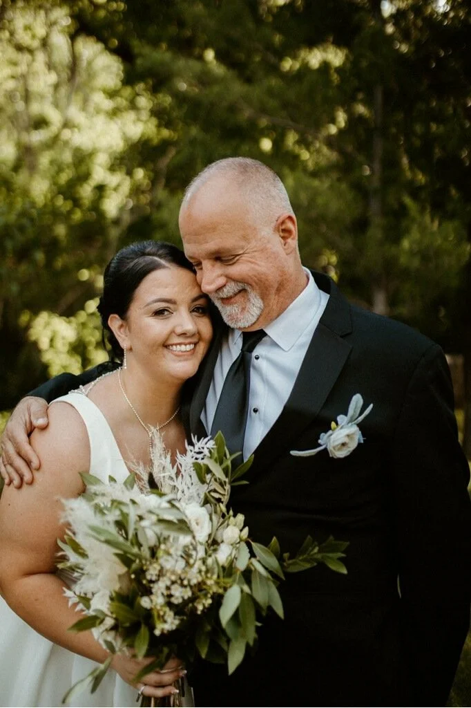 A bride and groom embrace outdoors during their wedding, smiling happily, with the bride holding a bouquet of white and green flowers and the groom wearing a suit with a boutonniere.