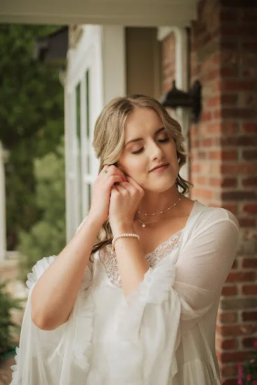 A young woman with blonde hair wearing a white dress, standing outside near a brick building with a white porch railing, holding her earring and smiling softly.