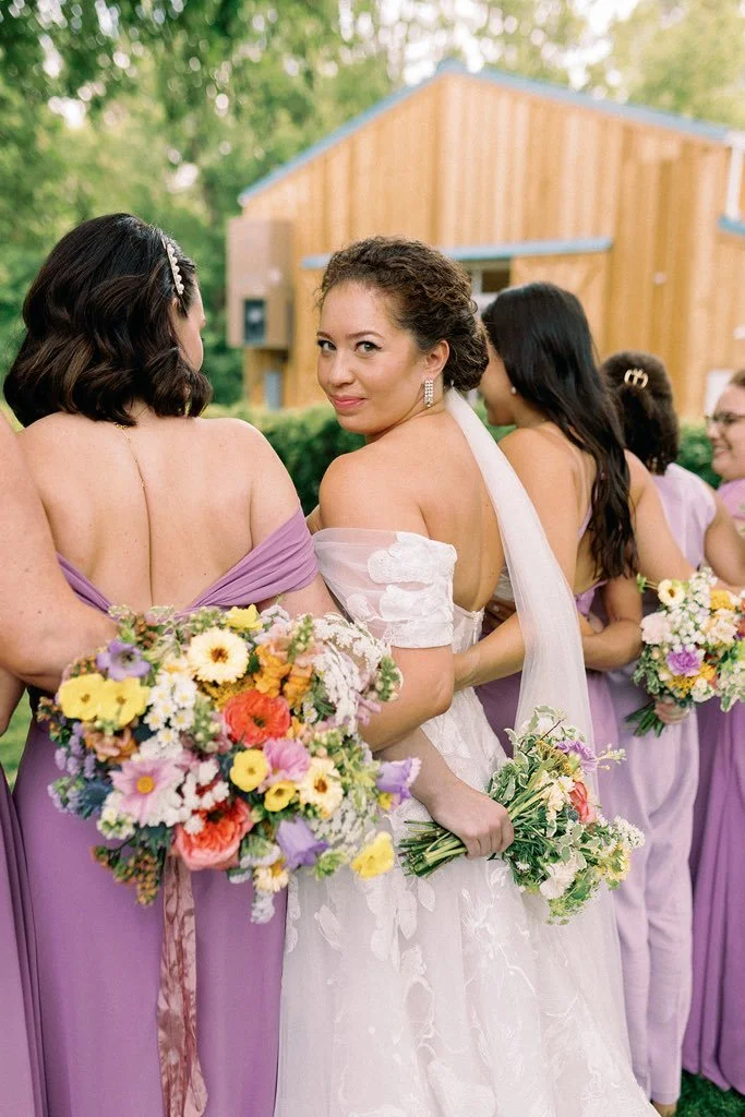 A bride in a white wedding dress holding a bouquet of flowers, surrounded by bridesmaids in lavender dresses at an outdoor wedding with a wooden building in the background.