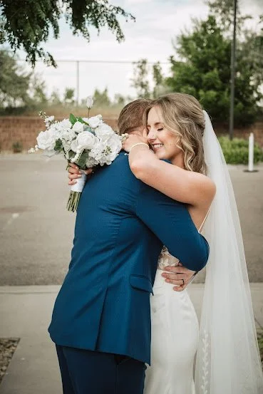 A bride and groom hugging outdoors on a wedding day. The bride is smiling and holding a bouquet of white flowers, and she is wearing a white wedding gown and veil. The groom is dressed in a blue suit.