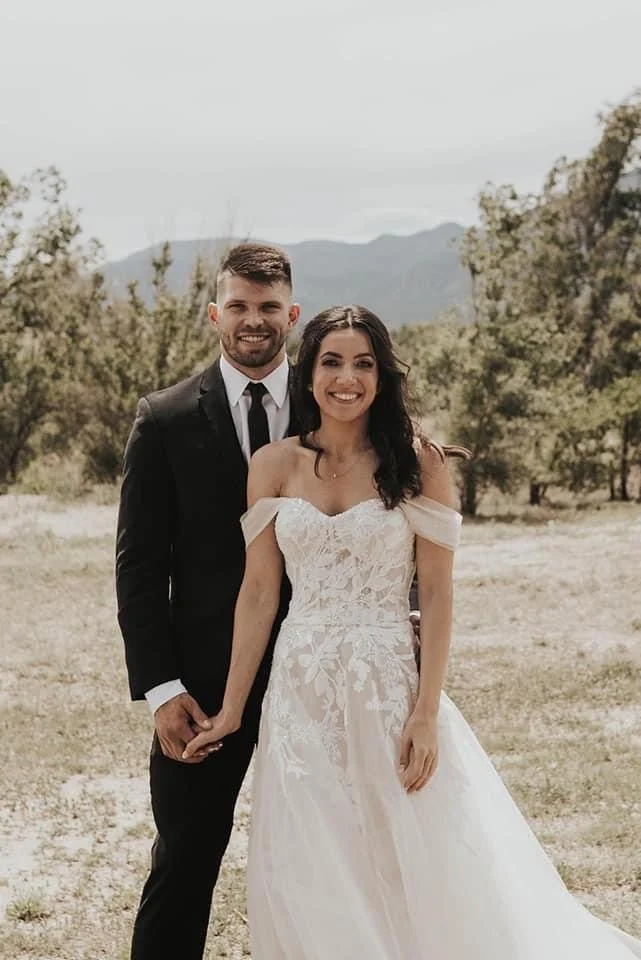 A newlywed couple holding hands outdoors, smiling, with a natural landscape of trees and mountains in the background.