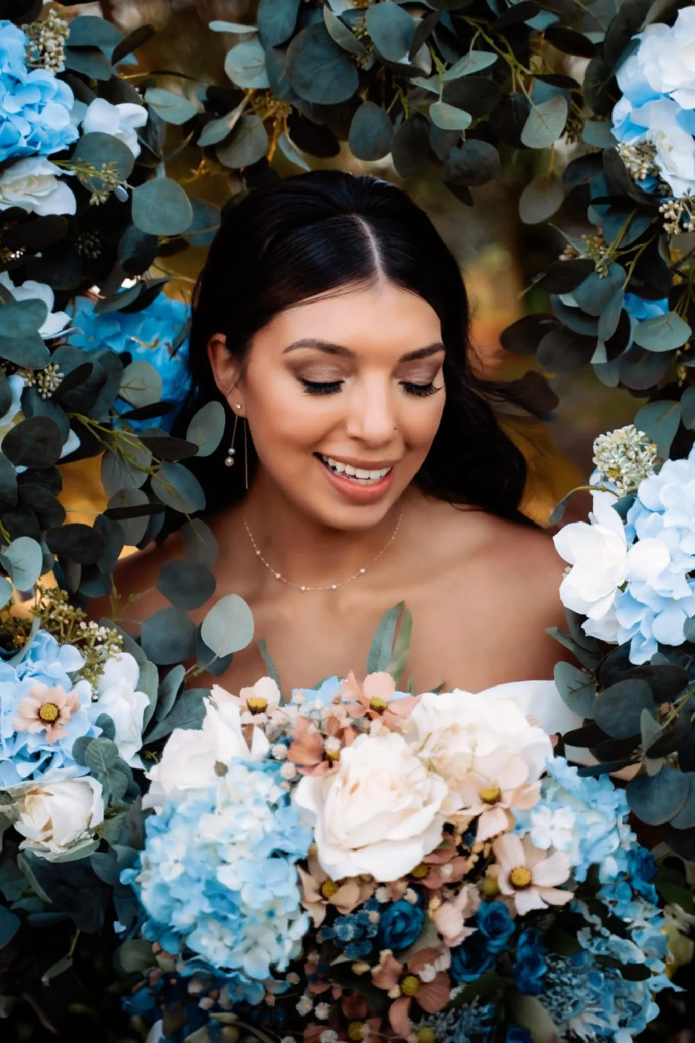 A woman with dark hair, smiling and looking down, surrounded by a lush arrangement of blue, white, and pale pink flowers, with green leaves.