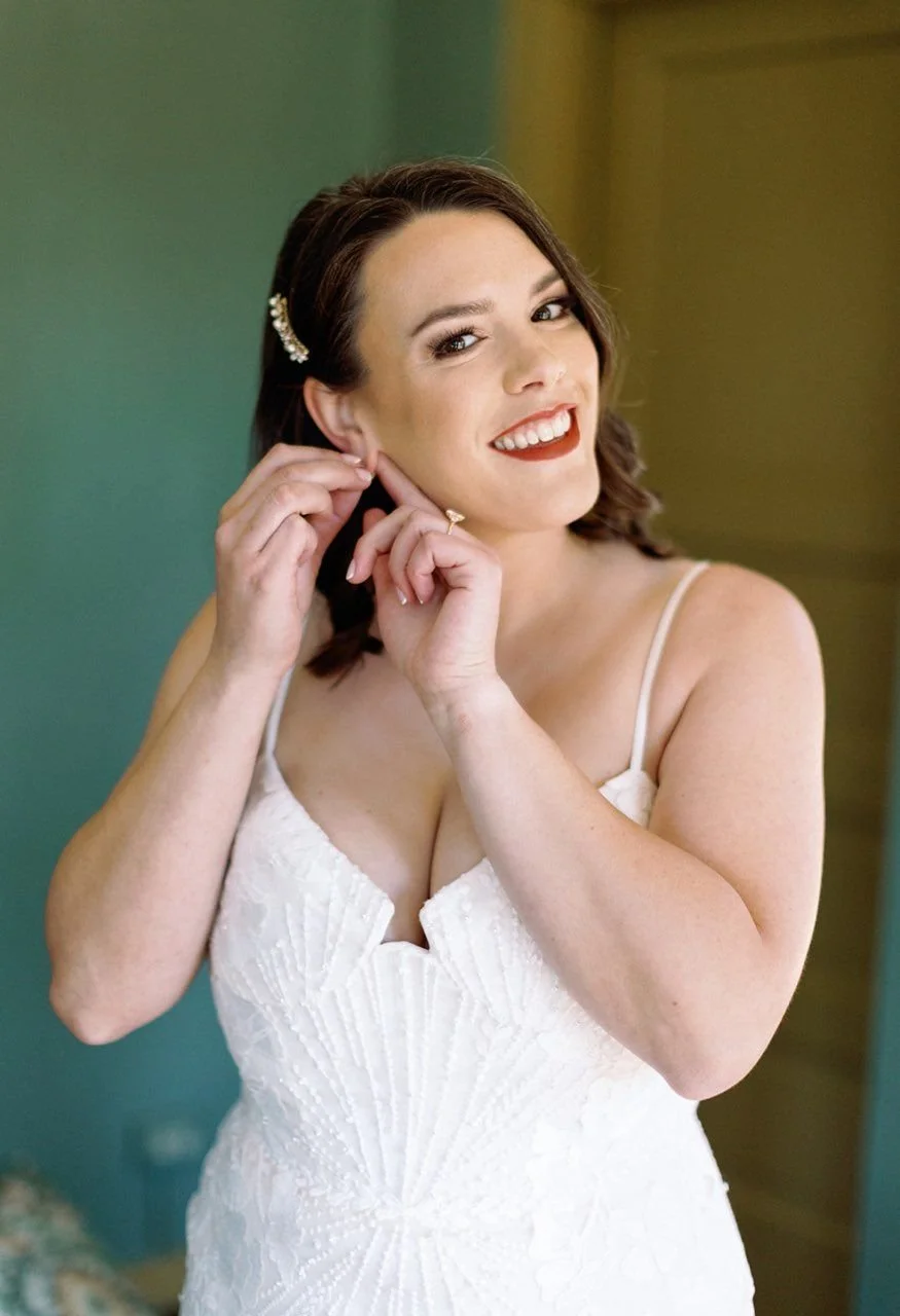 A smiling woman with dark hair wearing a white dress, putting on earrings.