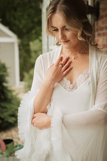 A woman in a white dress looking down with a gentle smile, standing outdoors in front of a garden and a house.