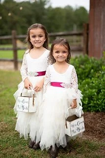 Two young girls in white dresses with pink sashes, holding small gift baskets, standing outdoors in a grassy area with a wooden fence in the background.