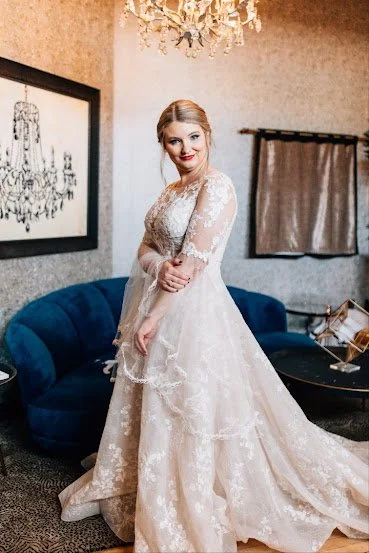A woman in a lace wedding gown standing in a living room with a chandelier, blue couch, framed artwork, and a curtain.