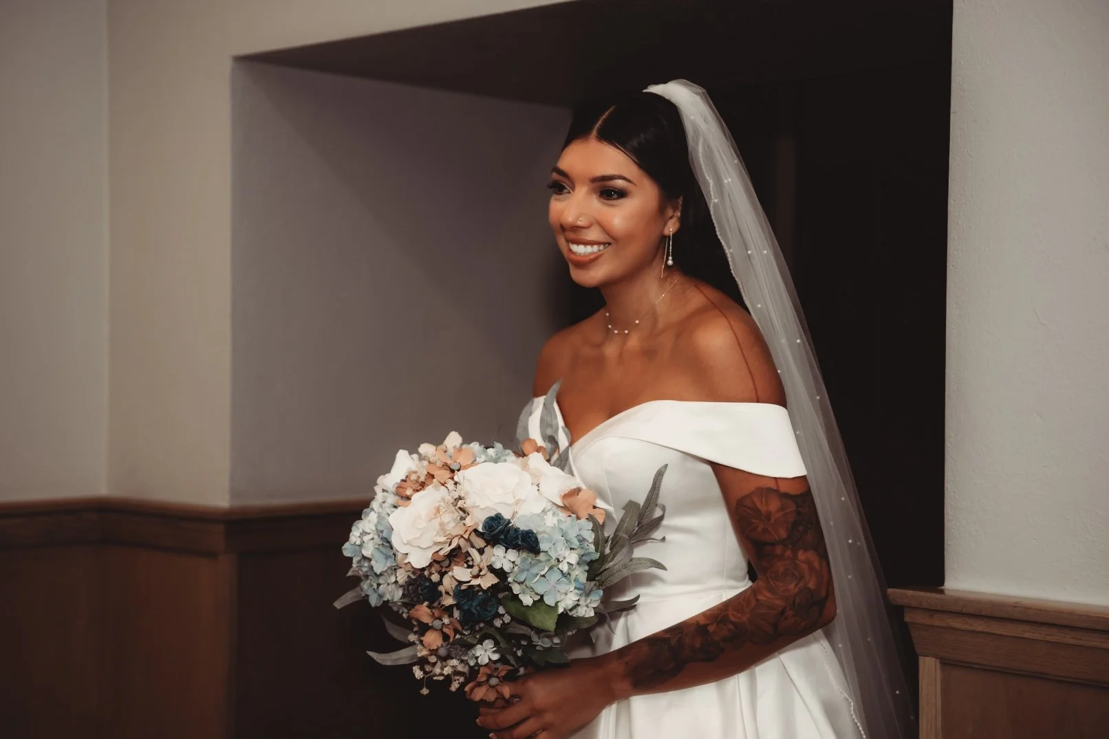 A smiling bride holding a bouquet of flowers, wearing an off-the-shoulder white wedding dress and veil.