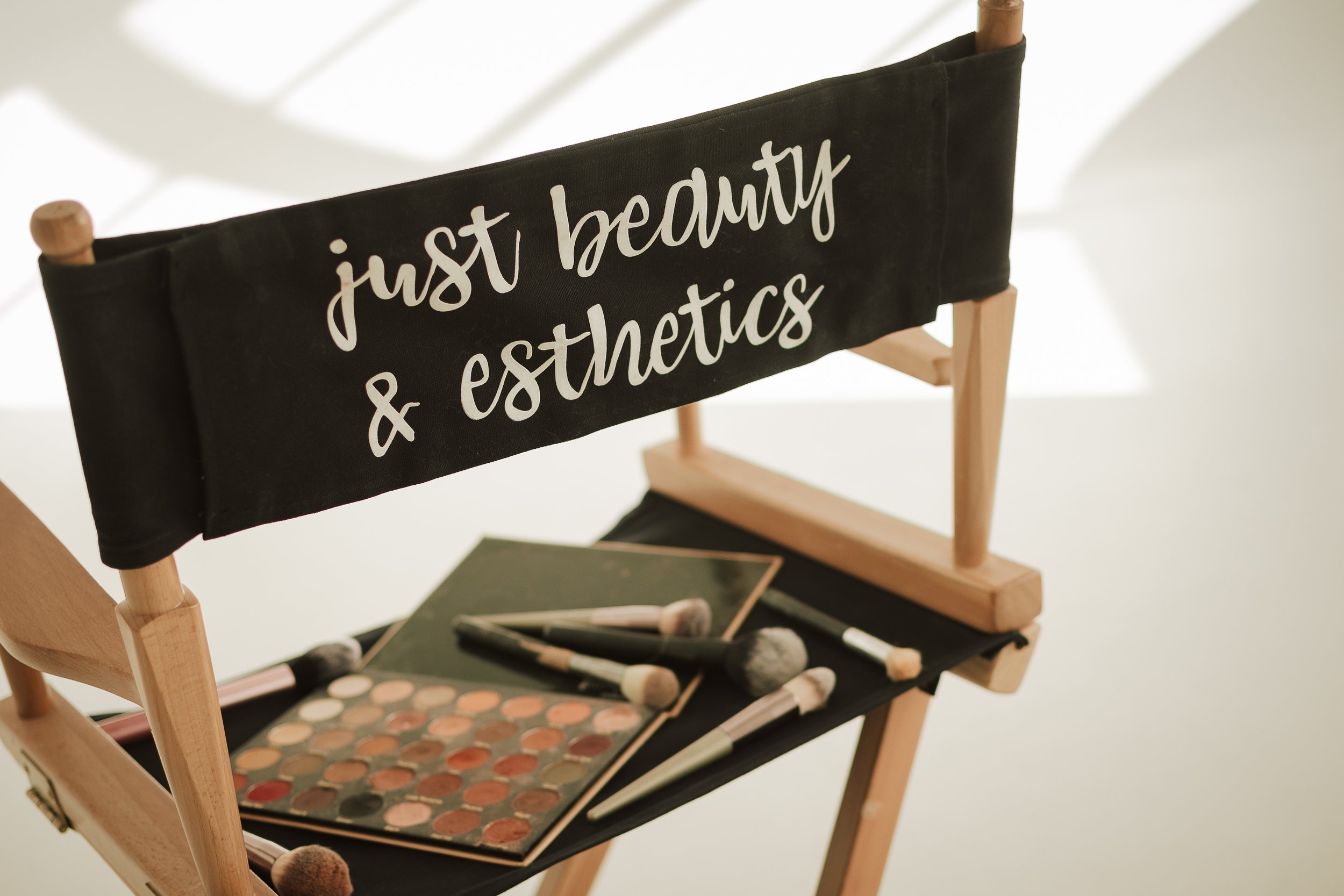 Makeup artist's chair with a black fabric backrest that says 'just beauty & esthetics' in white script, with makeup brushes and a palette of eyeshadows placed on a black seat.