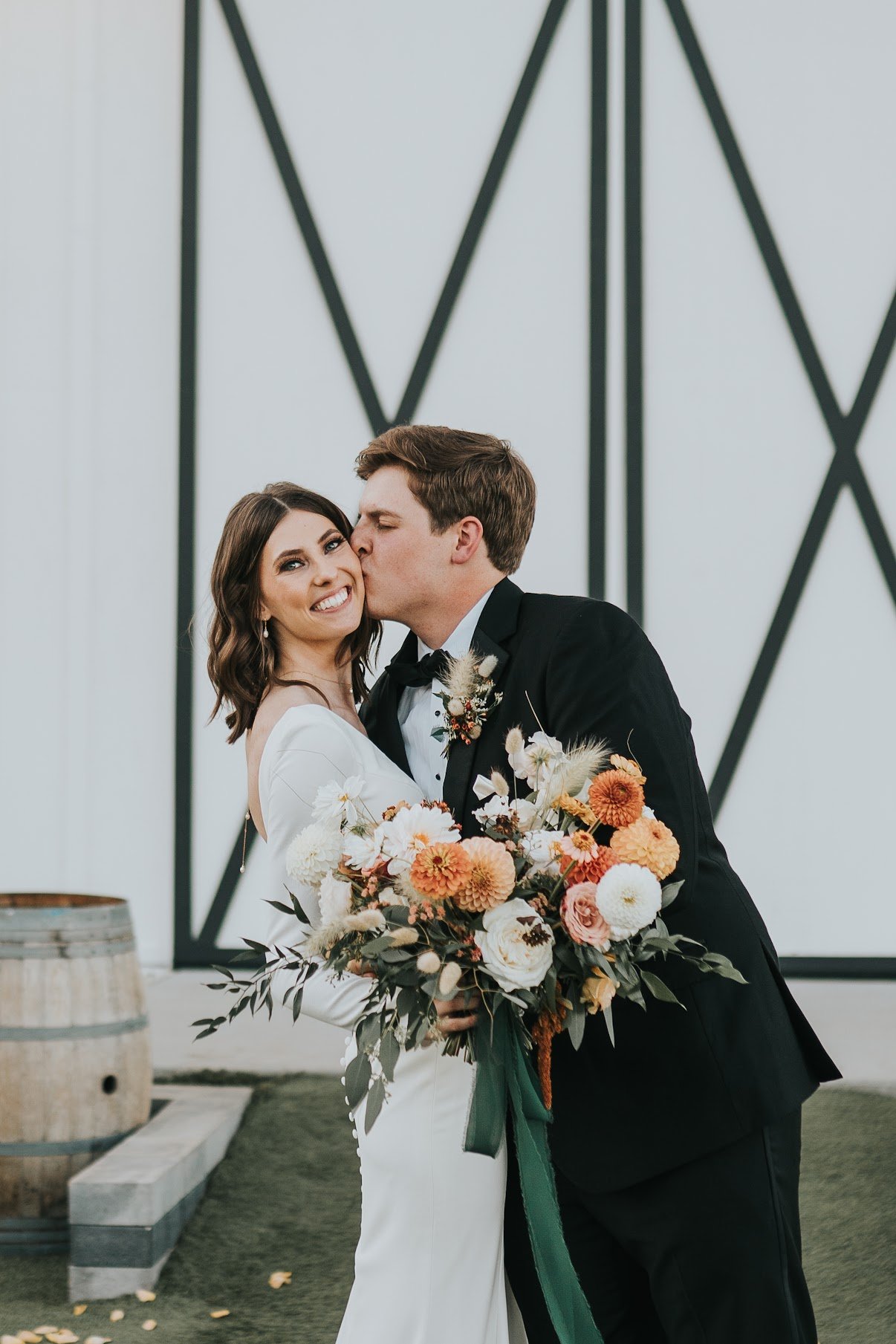 A newlywed couple sharing a kiss, with the groom kissing the bride on the cheek, holding a large bouquet of flowers. The bride is smiling at the camera, wearing a white wedding dress. The groom is in a black tuxedo. They are standing indoors with a m