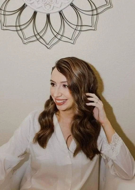 A woman with wavy brown hair smiling, wearing a white blouse with lace details on the sleeves, and touching her hair with her right hand, sitting in front of a wall with a decorative wire wall art resembling a lotus flower.