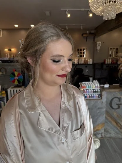 Young woman with styled hair and makeup wearing a shiny beige satin robe in a well-lit room with a chandelier and various decorations in the background.