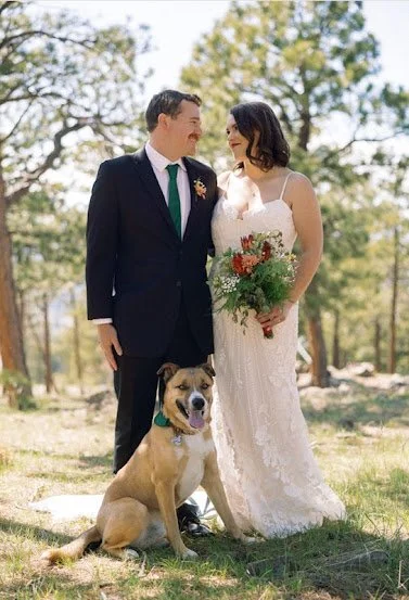 A newlywed couple standing outdoors in a park with their dog. The bride in a white lace wedding dress holds a bouquet, and the groom in a black suit with a green tie stands beside her. The dog is sitting in front of them, wearing a green collar, with