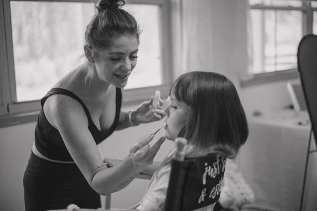 A woman applies makeup to a young girl sitting in a chair during a makeup session in a room with large windows.