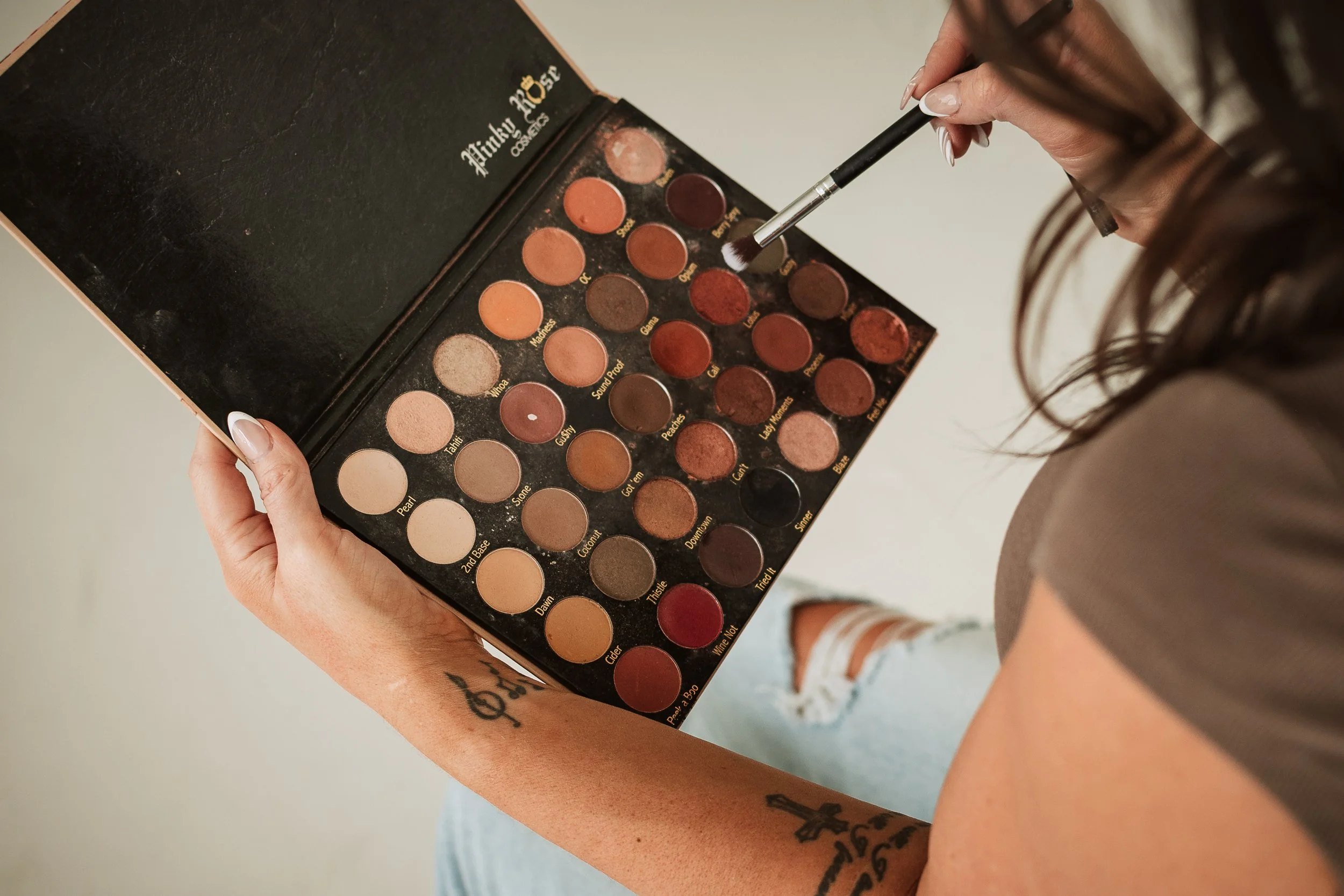 A woman holding an eyeshadow palette with various shades of nude, brown, and red eye shadows, and using a makeup brush to dip into one of the colors.