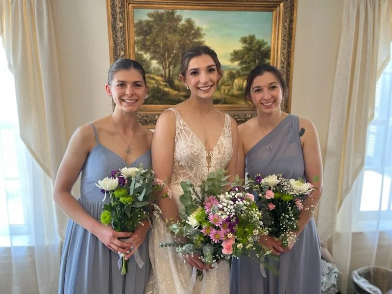 Three women in formal dresses holding bouquets of flowers, standing in a room with a landscape painting and curtained windows.