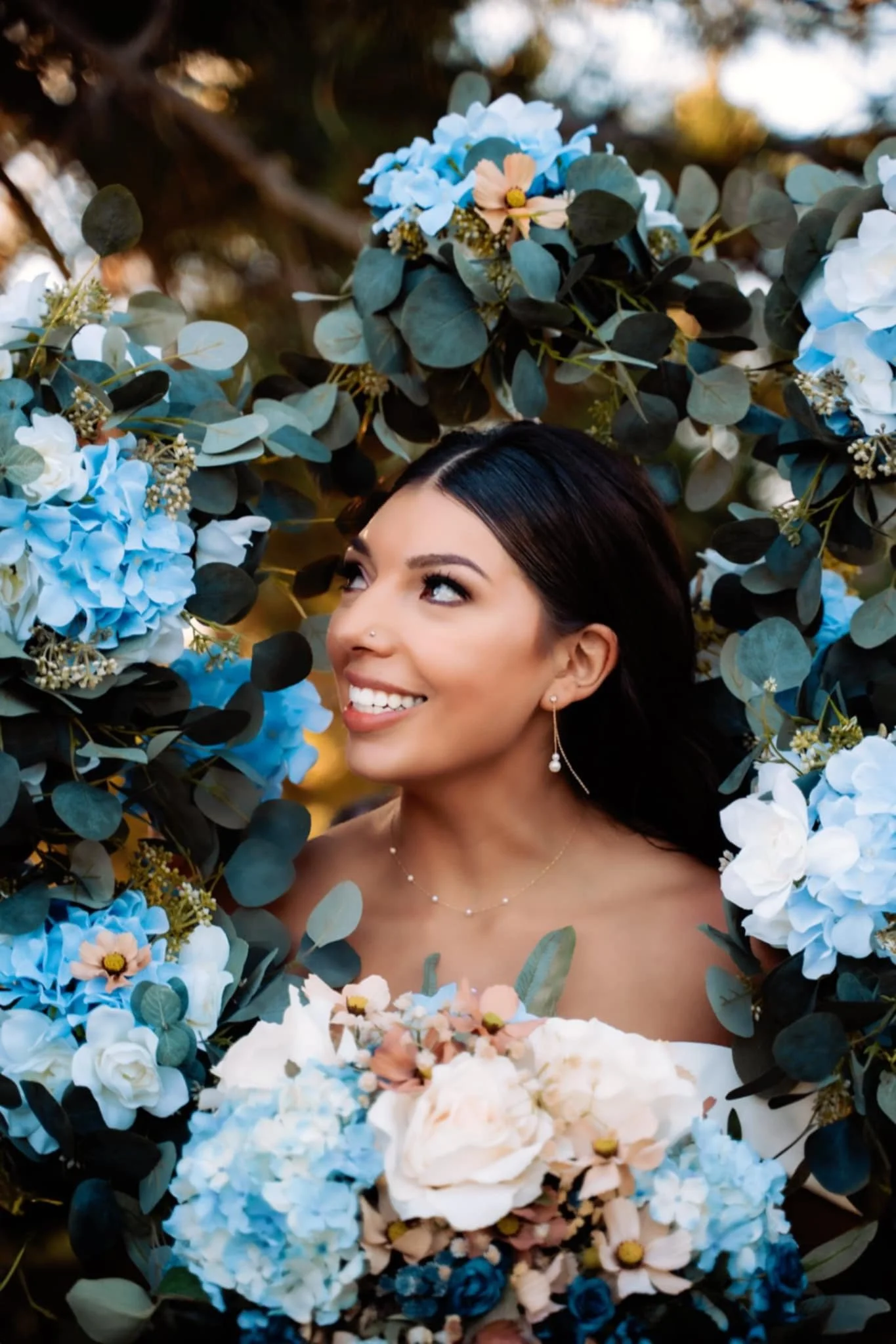 A woman smiling and looking upwards, surrounded by a circular arrangement of blue and white flowers with green leaves, outdoors during sunset.