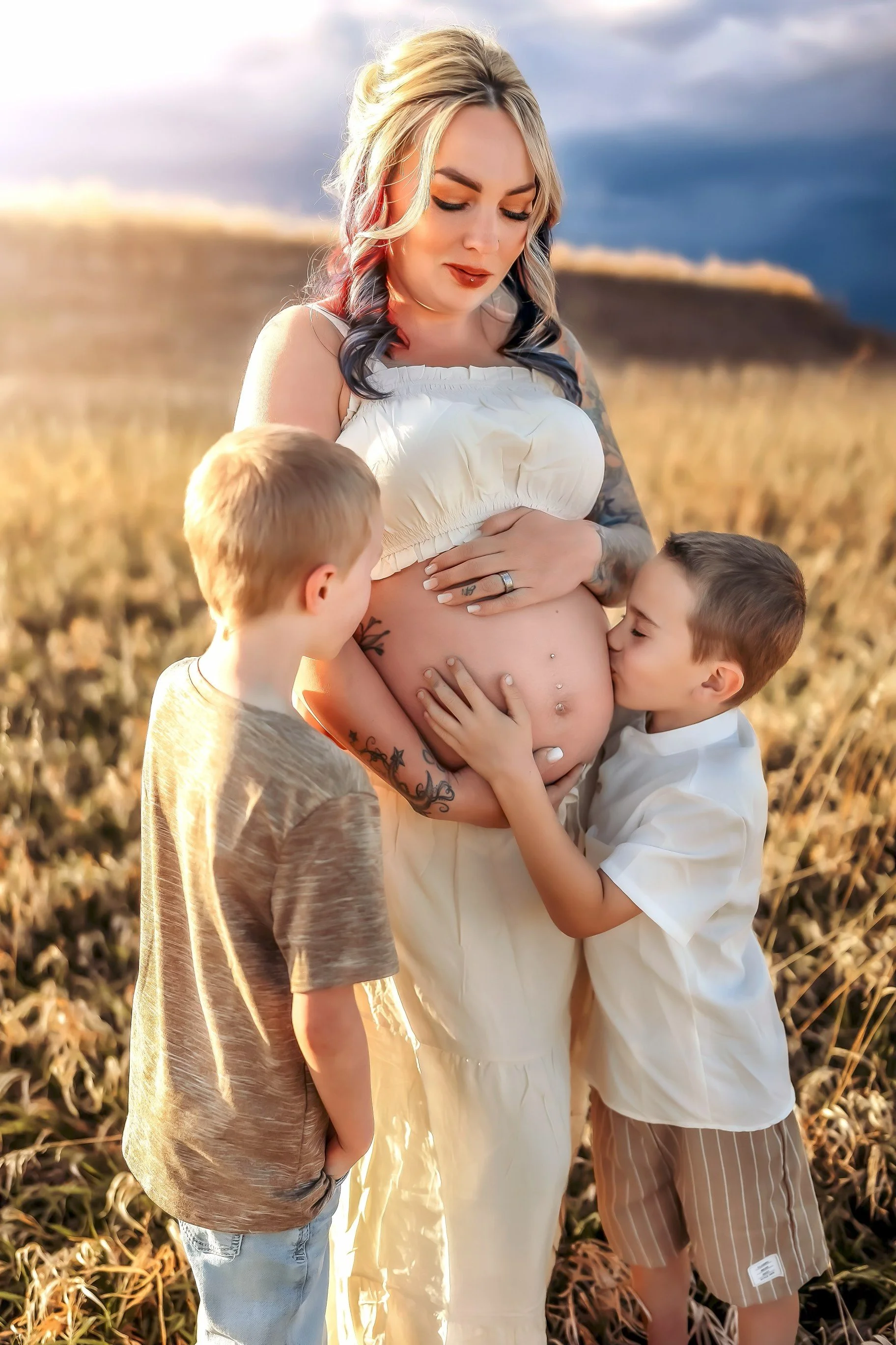 A pregnant woman with tattoos is outdoors in a field with two young boys kissing her belly.