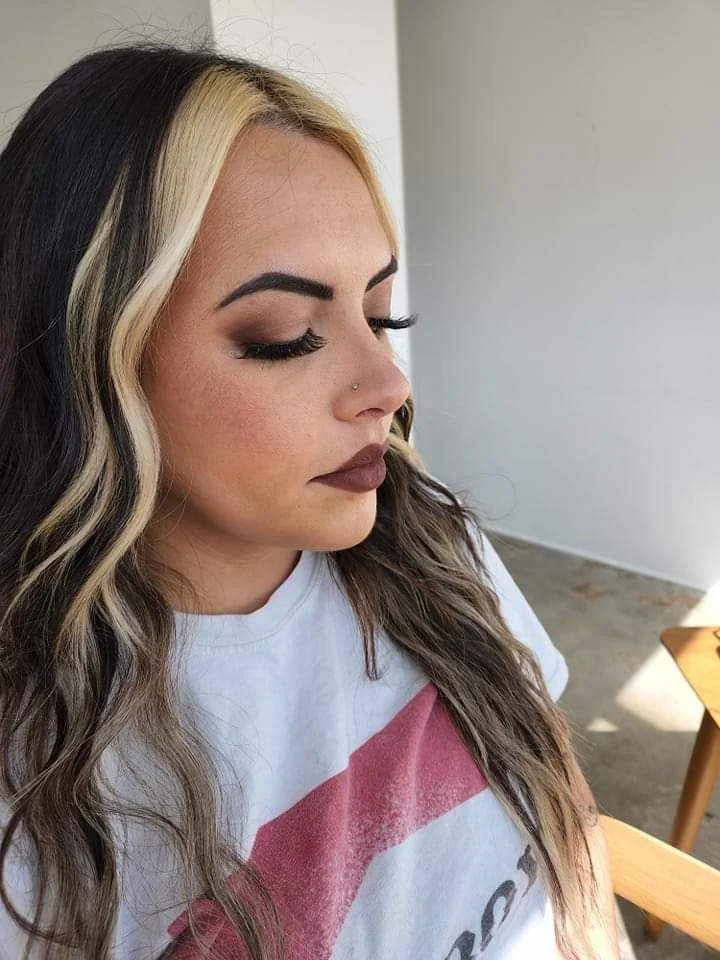 A woman with long wavy hair dyed black with blonde highlights, wearing makeup with dark lipstick, eye makeup, and a small nose piercing. She is wearing a white t-shirt with a red graphic and looking down, sitting near a wooden table.