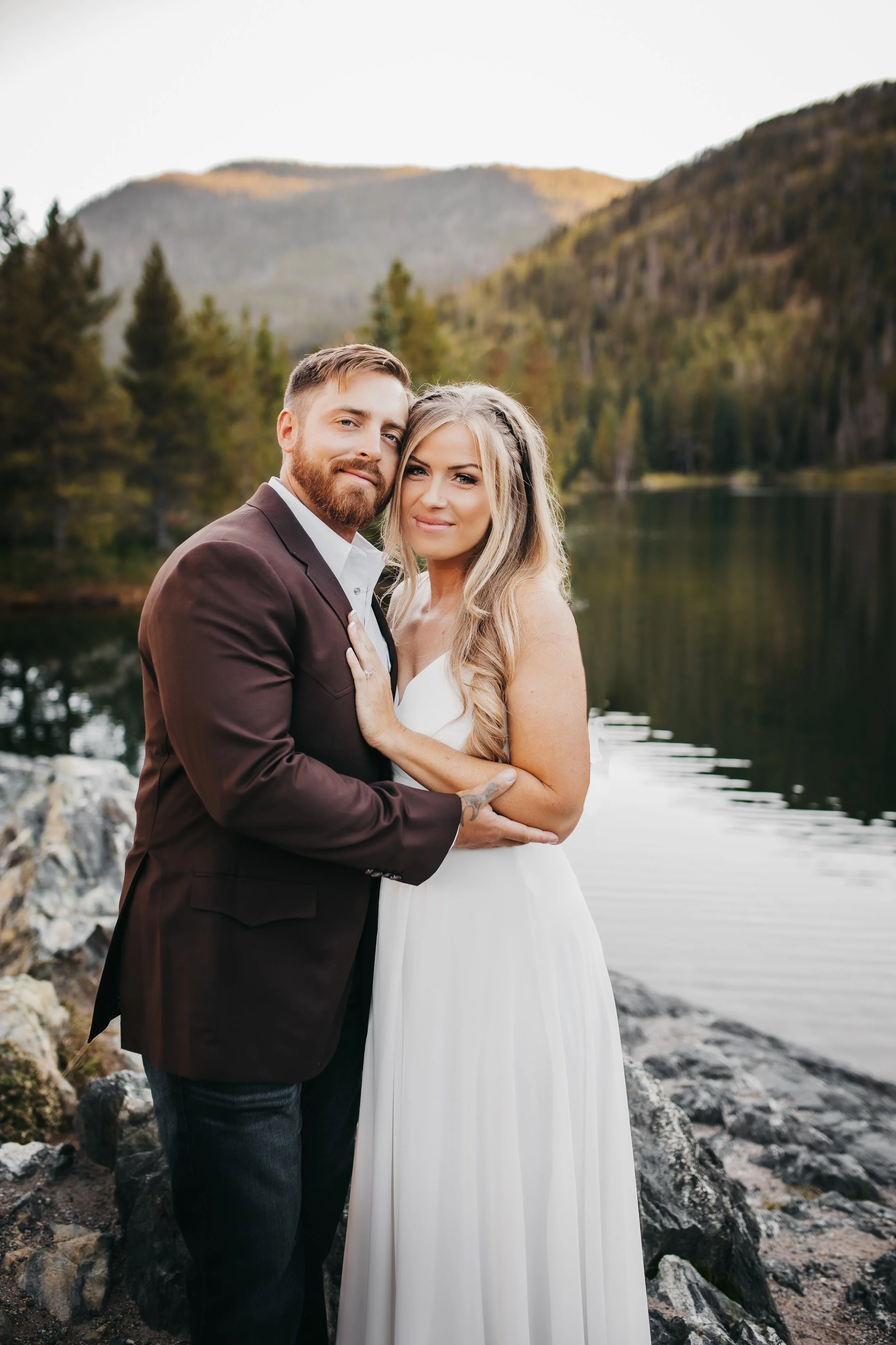 A bride and groom standing close together on rocky shore by a calm lake, with mountains and pine trees in the background during sunset.