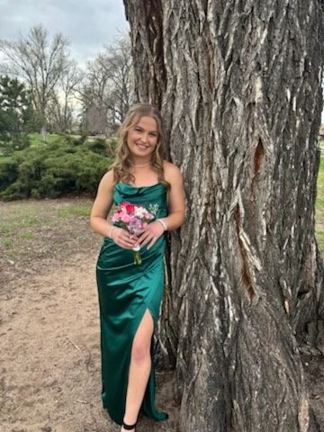 A young woman in a teal dress holding a small bouquet of flowers, standing next to a large tree outdoors.