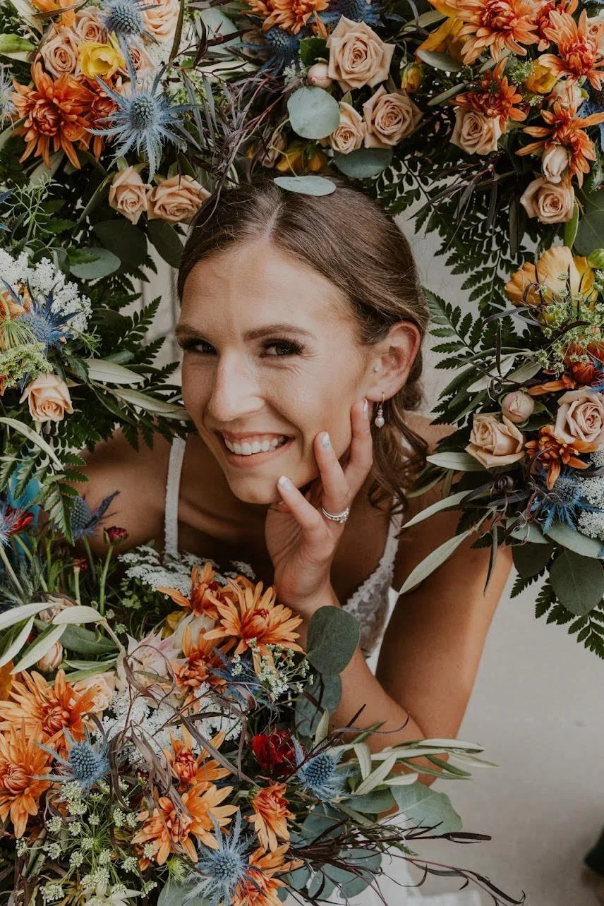 A woman smiling and winking through a floral wreath made of various orange, peach, blue, white, and green flowers and foliage.