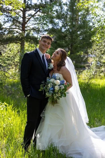 A bride and groom standing outdoors on a sunny day in a green, wooded area, with the bride holding a bouquet of white and blue flowers.