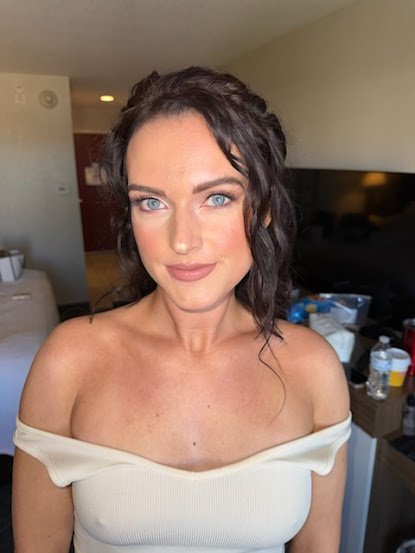 A woman with dark curly hair and blue eyes, wearing an off-the-shoulder beige top, posing indoors with a living room background.