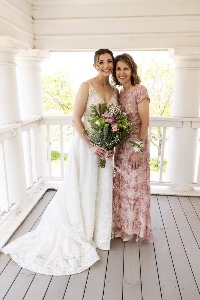 Two women standing on a porch, one in a wedding dress holding a bouquet of flowers, and the other in a pink lace dress, smiling for a photo.
