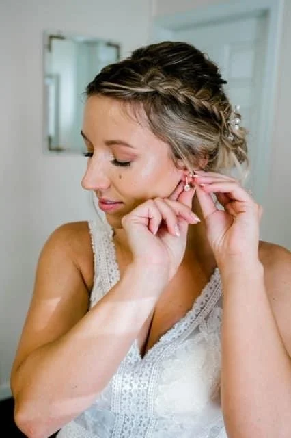 A woman with styled hair putting on pearl earrings in a bright room.