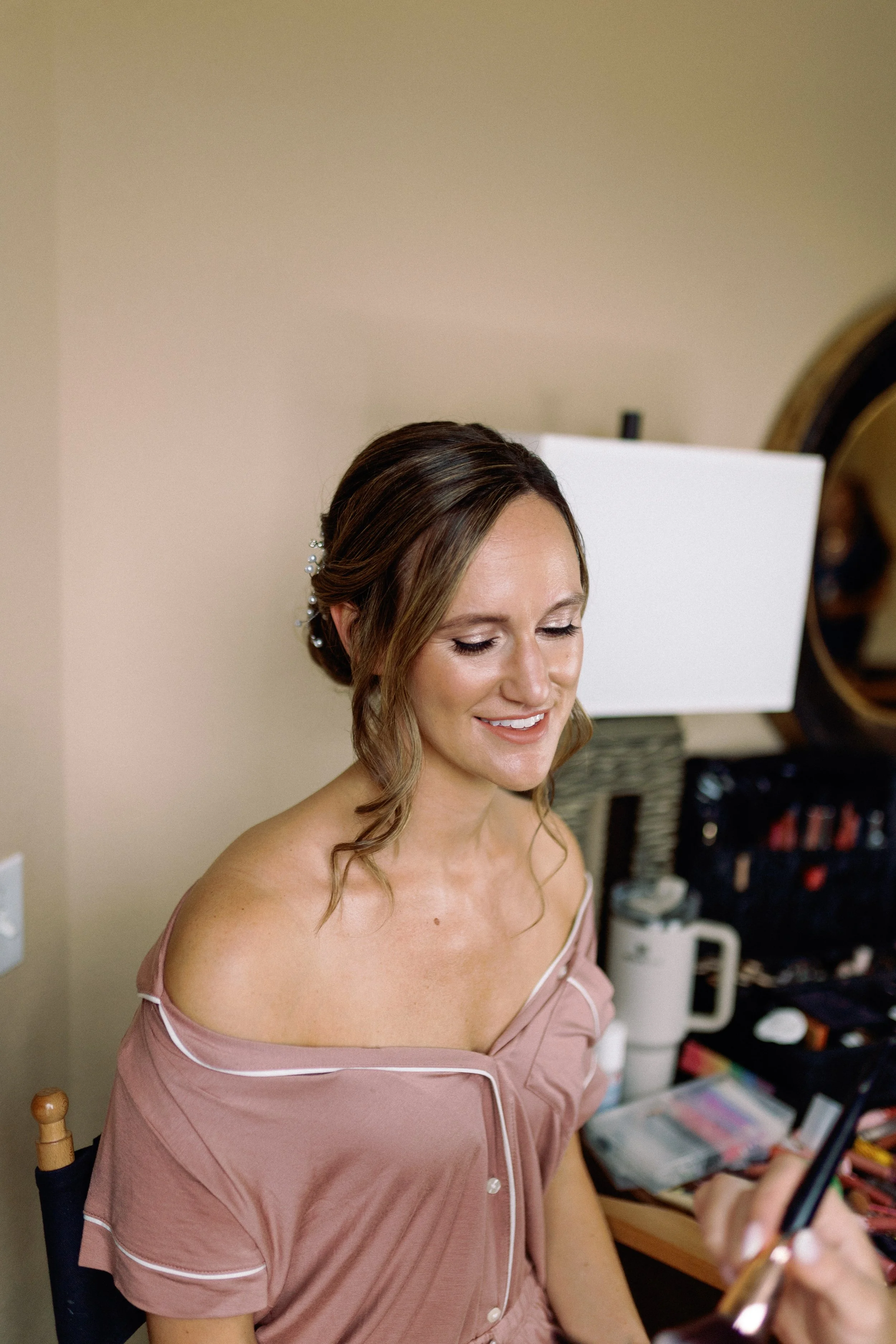 A woman with a side braid and a decorative hairpin is getting her makeup done, seated in front of a mirror and makeup supplies.