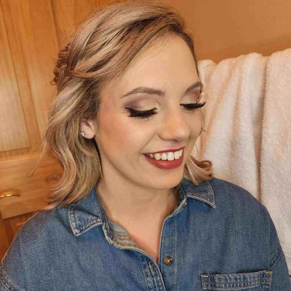 A woman with blonde hair styled in loose waves, wearing makeup, earrings, a denim shirt, and smiling with her eyes closed.
