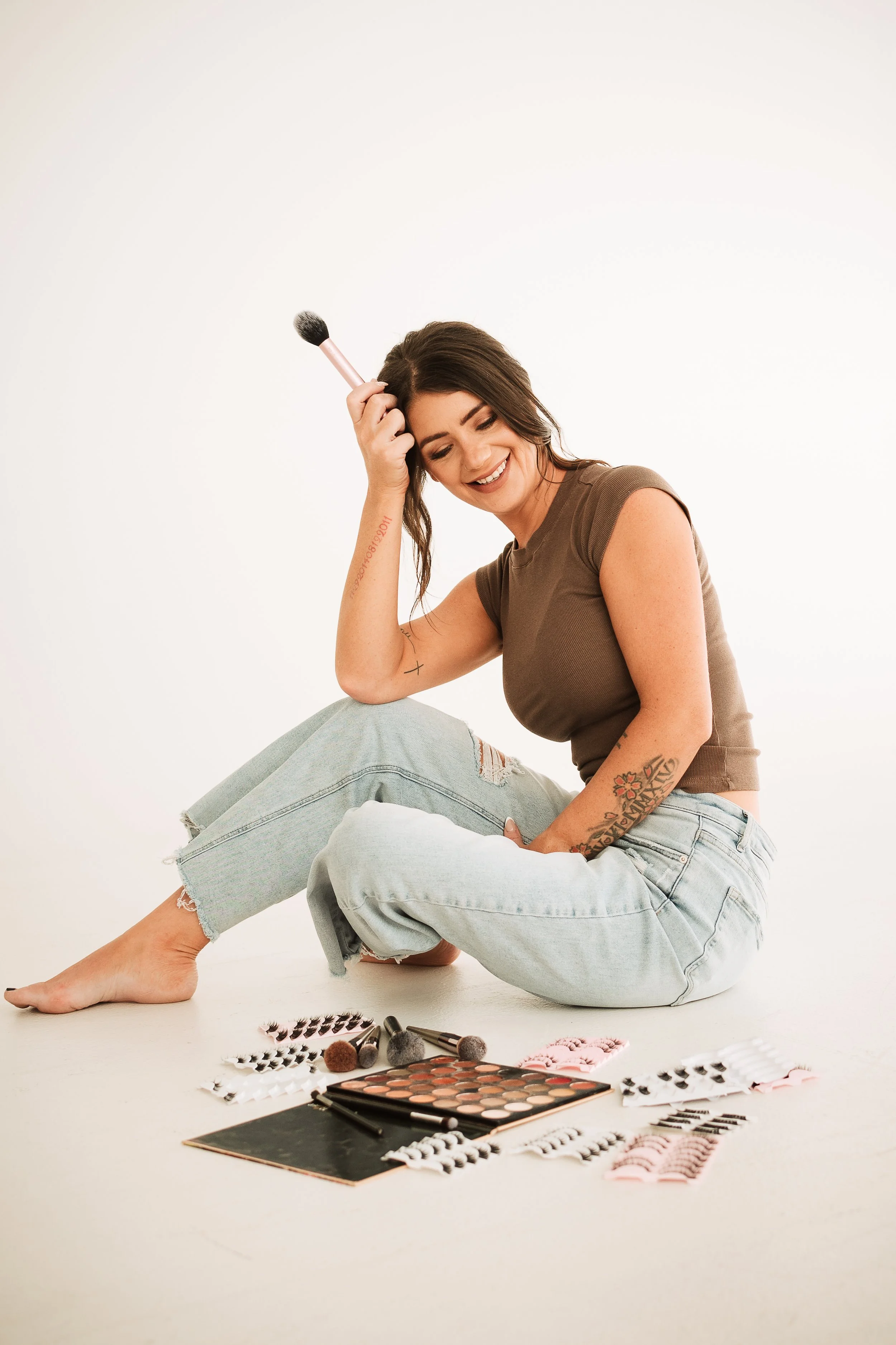 Young woman sitting on the floor with makeup products around her, smiling and holding a makeup brush to her hair.