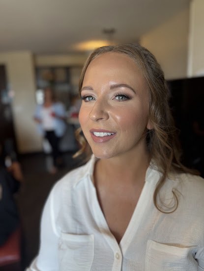 A young woman with wavy light brown hair and makeup, wearing a white blouse, smiling indoors with blurred people in the background.