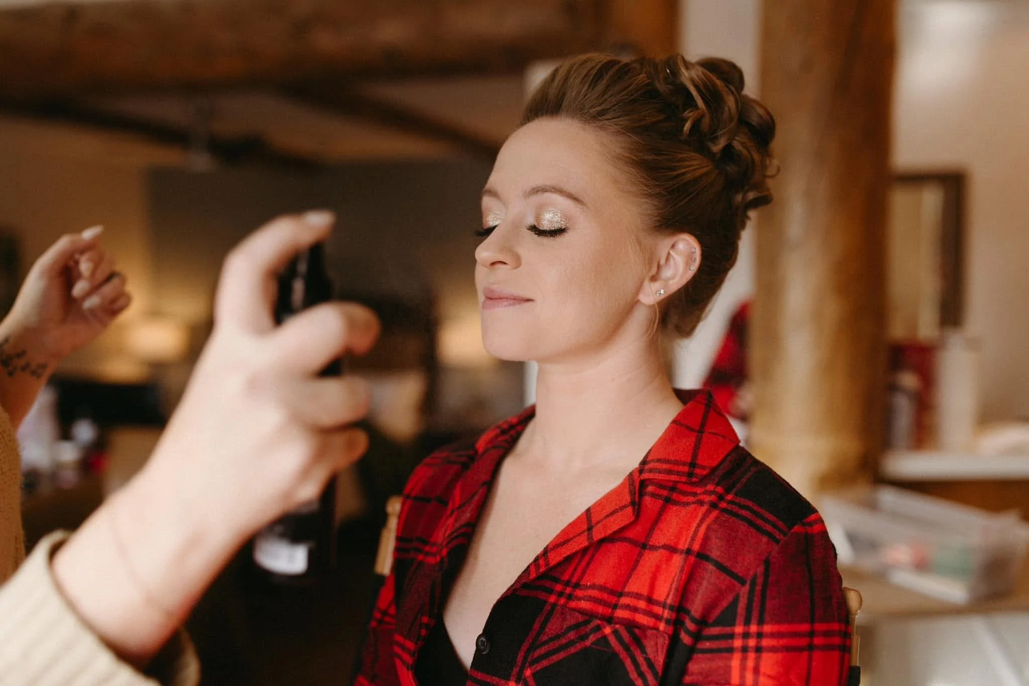A woman with a styled updo hairstyle getting her makeup done, wearing a red and black plaid shirt, with closed eyes and a content expression.