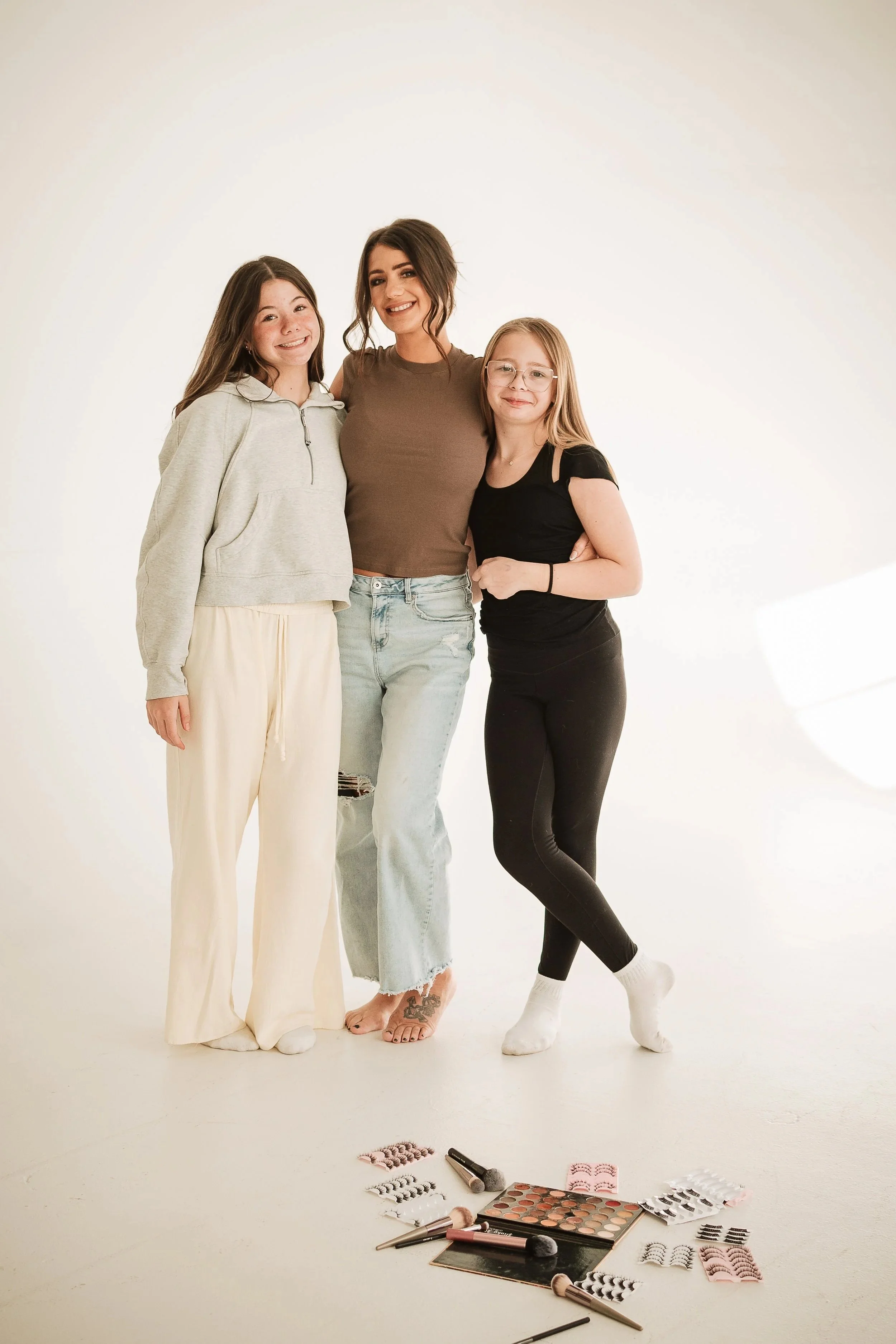 A woman and two girls standing together in a photography studio, smiling at the camera. There is makeup and beauty products laid out on the floor in front of them.