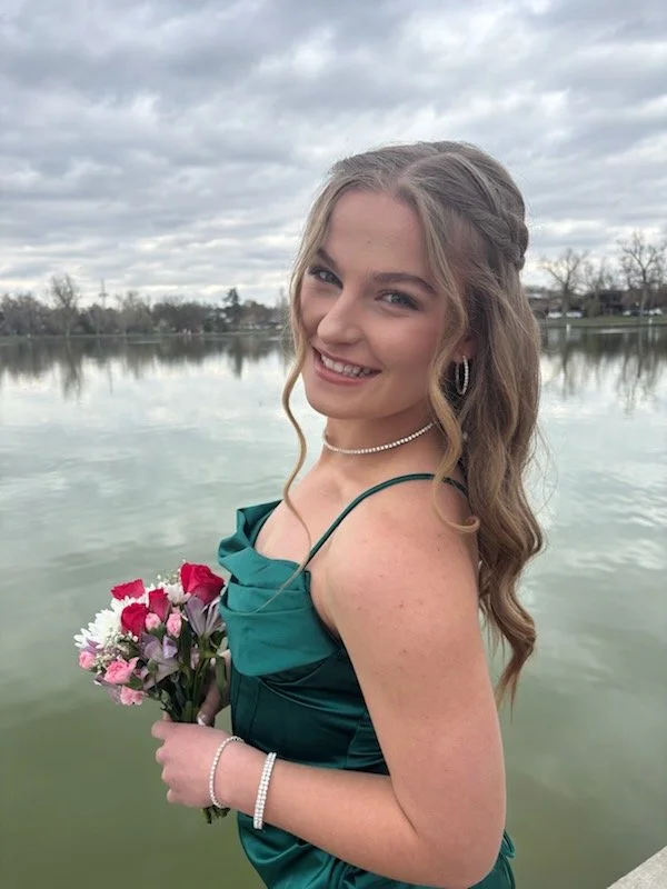 A young woman with long wavy blonde hair, wearing a teal dress, holding a bouquet of pink, red, and white flowers, standing by a body of water with trees and buildings in the background, under an overcast sky.