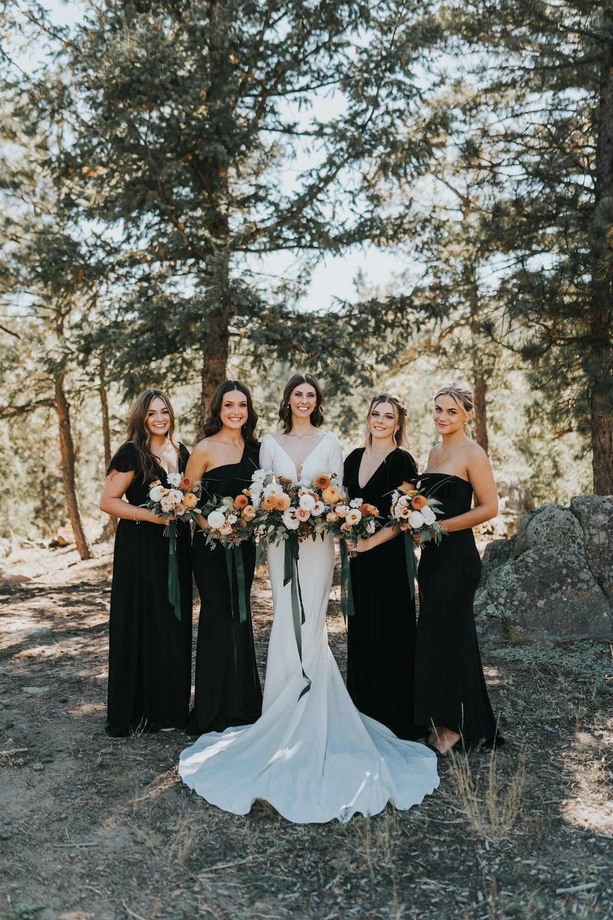A bride in a white wedding dress standing in the center with four bridesmaids in black dresses on a forested trail, all holding bouquets of flowers.