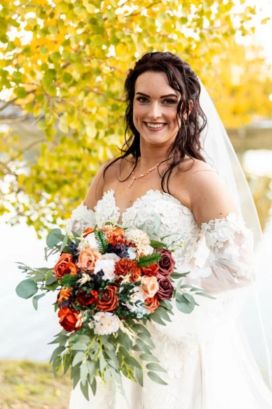 smiling bride in a white lace wedding dress holding a vibrant bouquet of flowers outdoors with yellow autumn leaves in the background