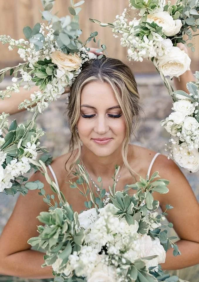 A woman with blonde hair and makeup is smiling with her eyes closed, surrounded by an arch of white and pale pink flowers and green foliage.