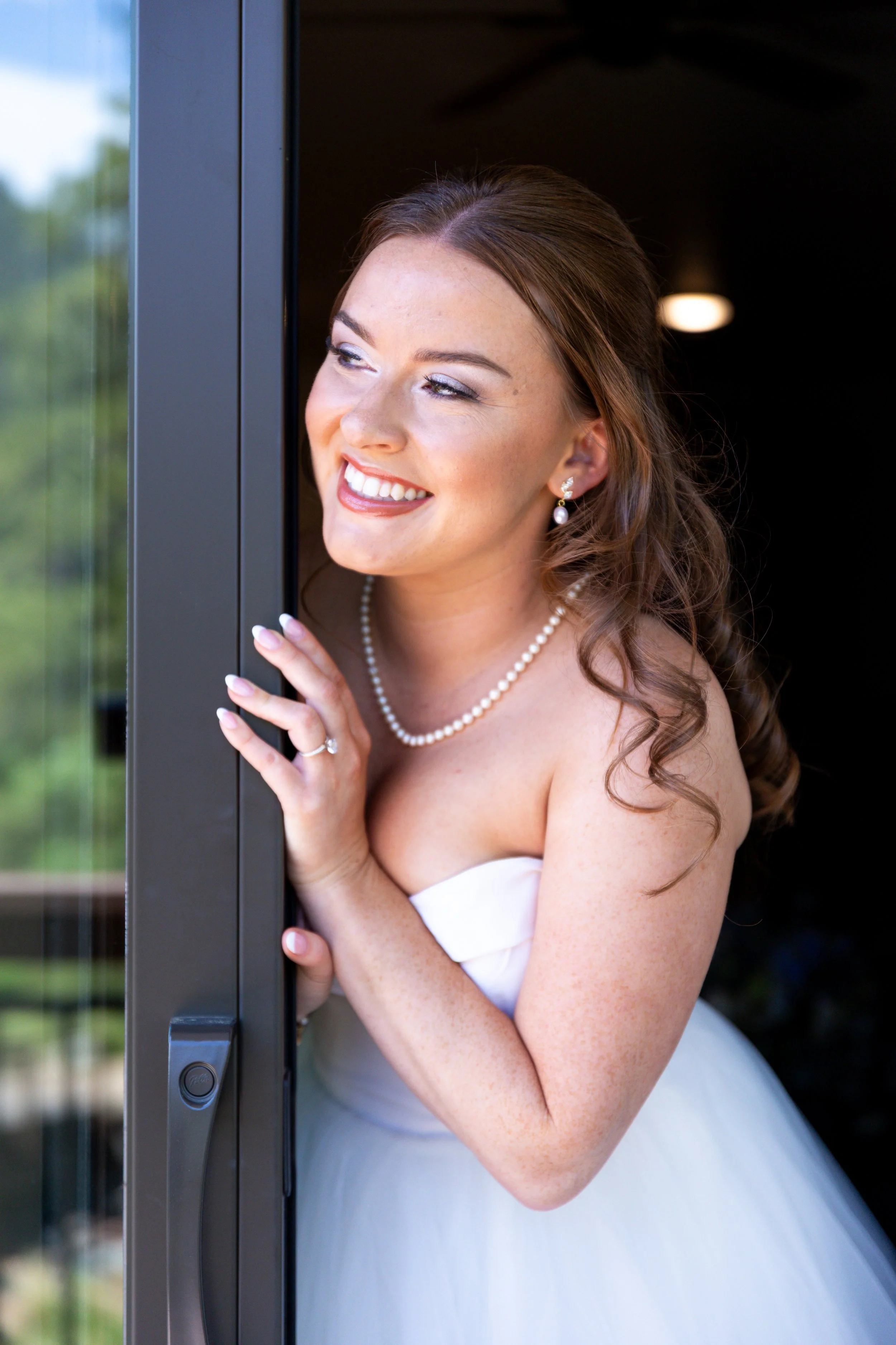 A smiling woman in a white wedding dress with pearl jewelry peering out of a doorway.