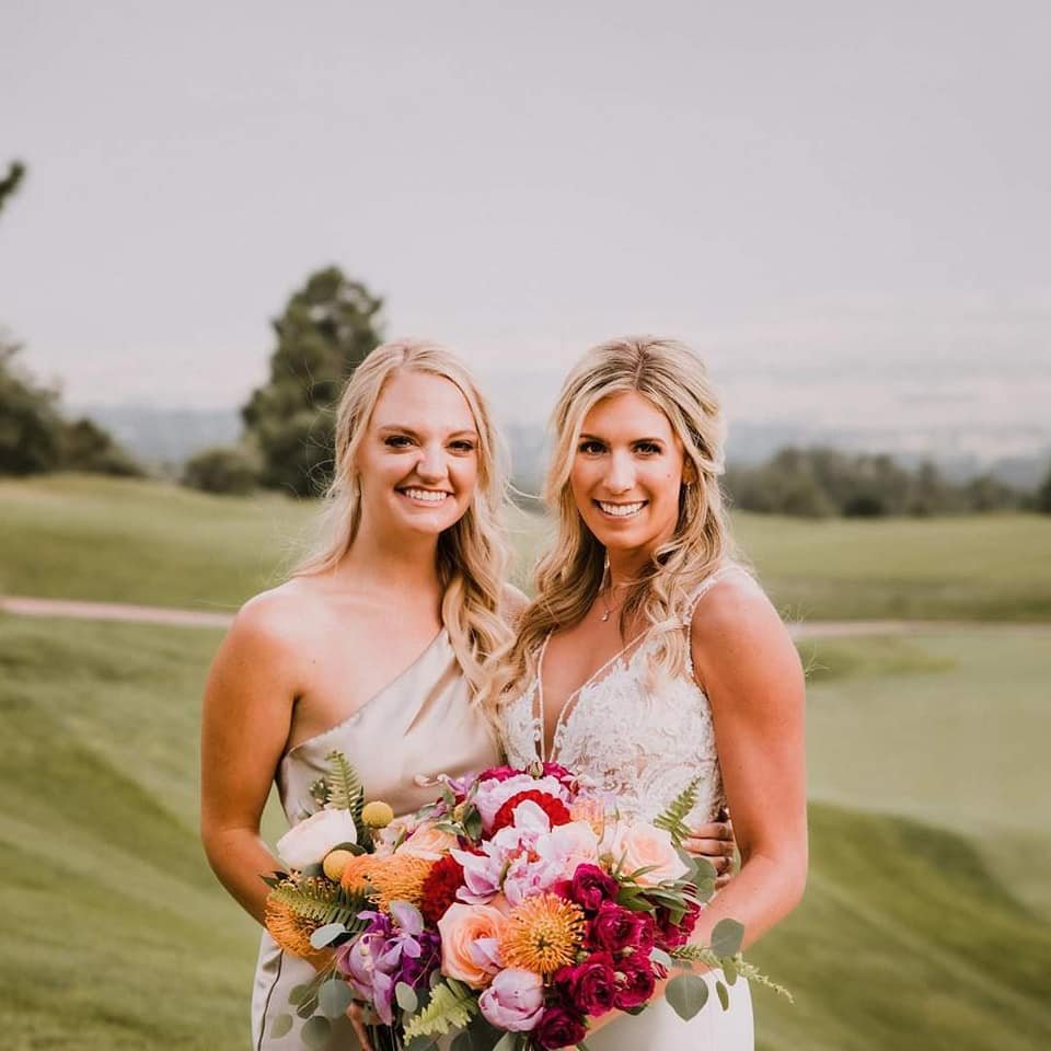 Two women, dressed in wedding attire, smiling and standing outdoors on a grassy field with trees in the background, one holding a colorful bouquet of flowers.