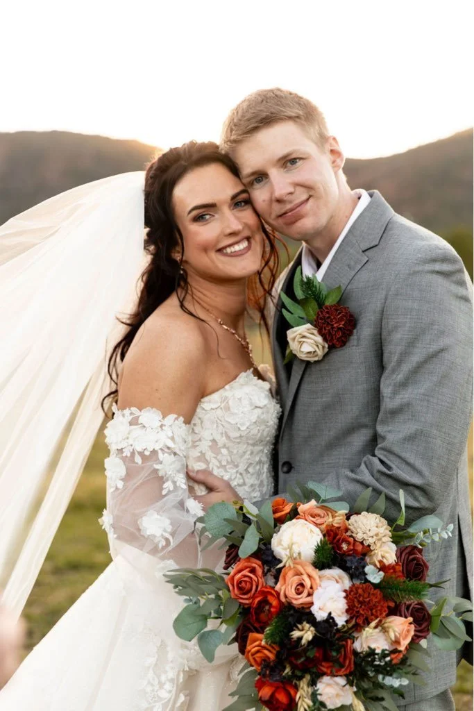 A bride and groom smiling at the camera, embracing outdoors at sunset. The bride wears a lace wedding dress with off-shoulder sleeves and a veil, holding a large bouquet of red, white, and peach flowers with greenery. The groom wears a gray suit with